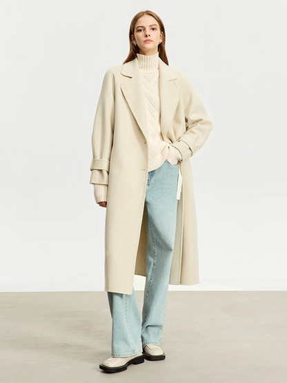 A woman wearing a turtle neck knit sweater with a tie design on the side in a cream color and a pair of denim jeans and an oversized trench coat.