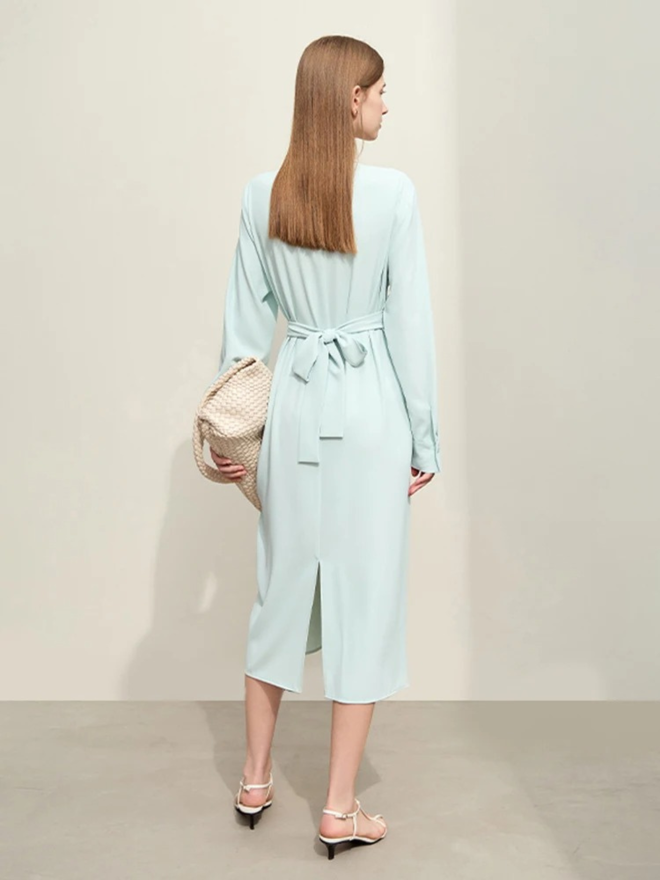 A woman turned away wearing a powder blue, long sleeved, midi dress with a tie design to cinch the waist and a slit at the back for ease of movement. 