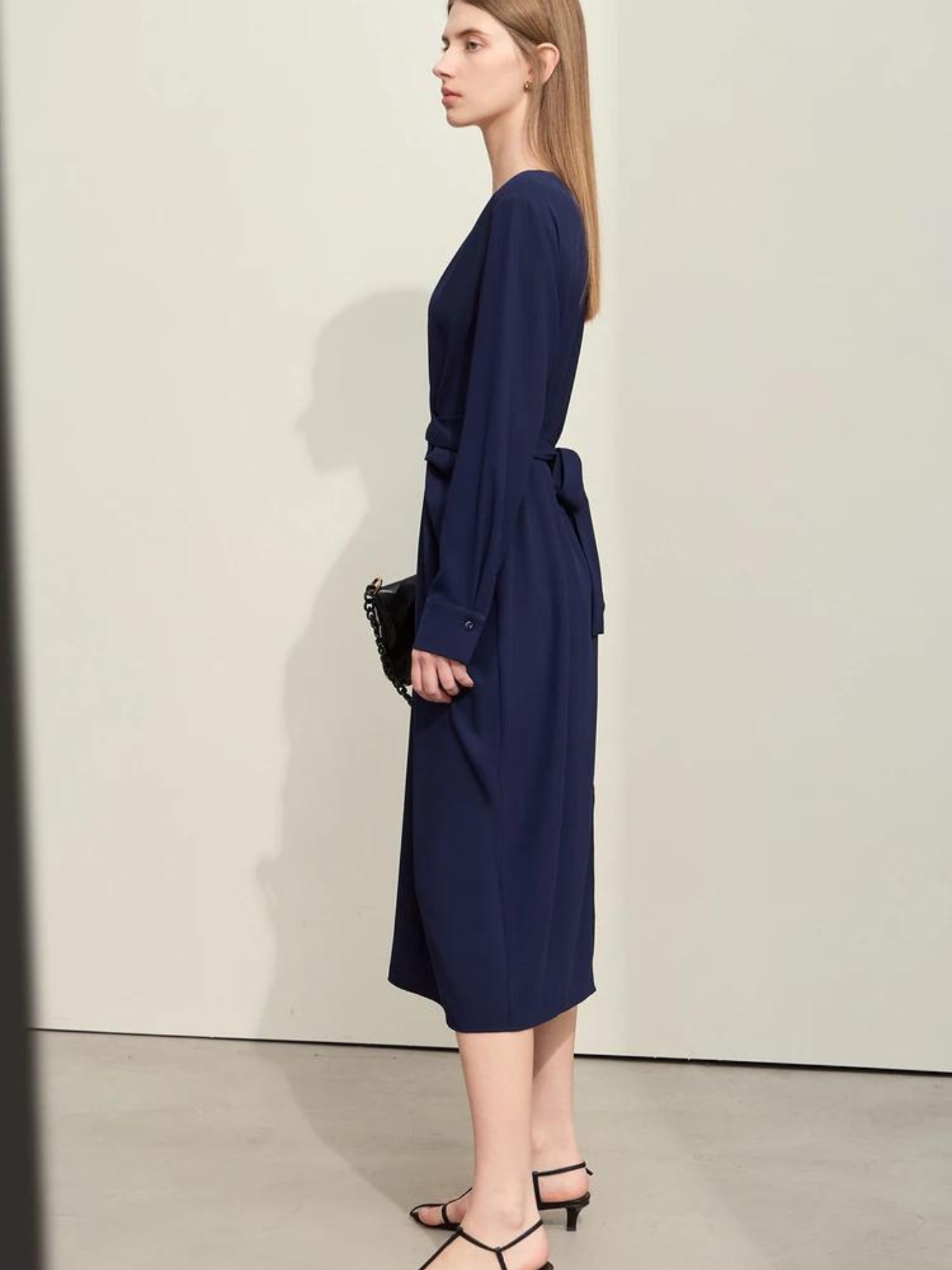 A stylish woman wearing a navy blue, long sleeved, midi dress with a ruching and tie design to cinch the waist,  and a slit at the back for ease of movement. She paired it with a black bag and black sandals for a monochrome look.