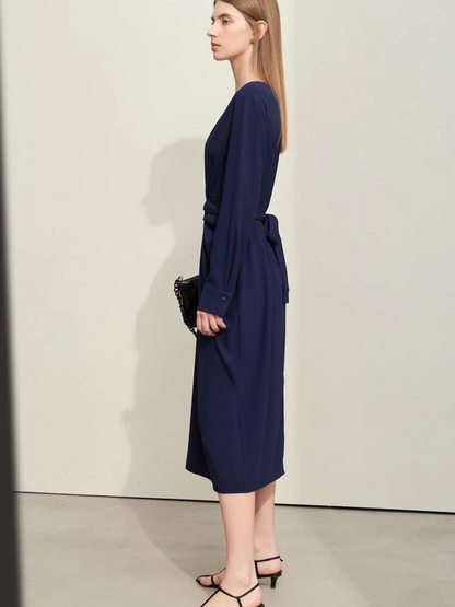 A stylish woman wearing a navy blue, long sleeved, midi dress with a ruching and tie design to cinch the waist,  and a slit at the back for ease of movement. She paired it with a black bag and black sandals for a monochrome look.