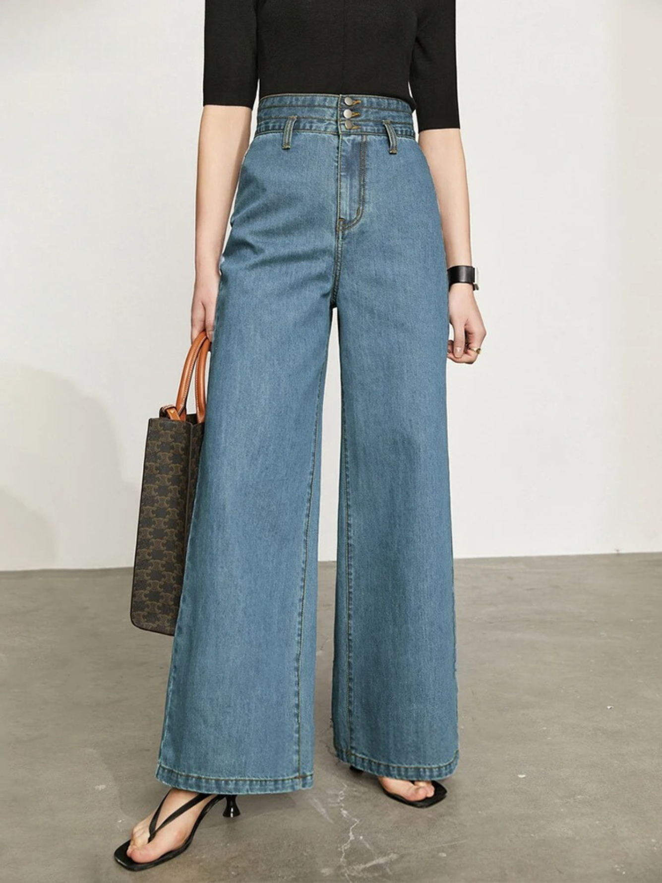 A woman wearing a fitted black shirt with a pair of retro-inspired, wide leg jeans with a cinched waist design and a pair of sandals.