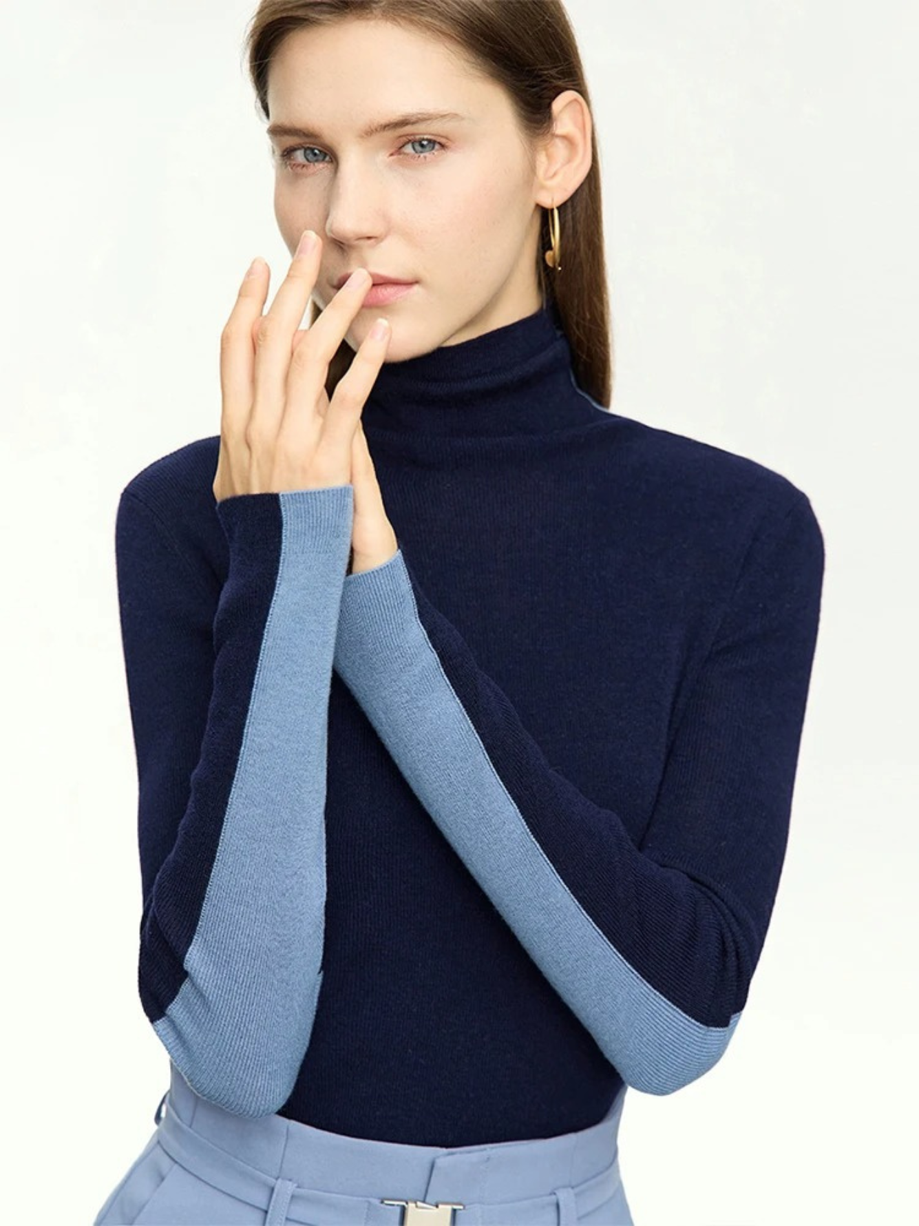 A woman wearing a mock neck, dual toned sweater in light and dark blue with a pair of light blue belted high waisted pants with a belt detail.